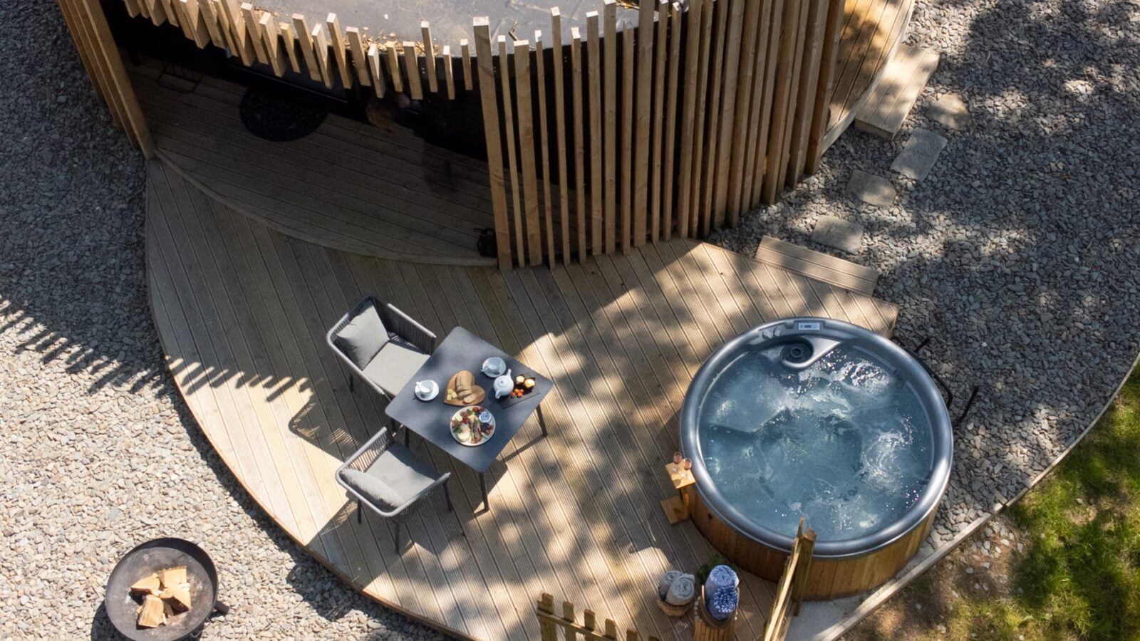 Aerial view of a circular patio at Pond View Lodges, featuring a hot tub and small dining area. Surrounded by a curved wooden screen, gravel, and grassy area, the table is set with plates and bowls. A cozy fire pit completes this serene setting.