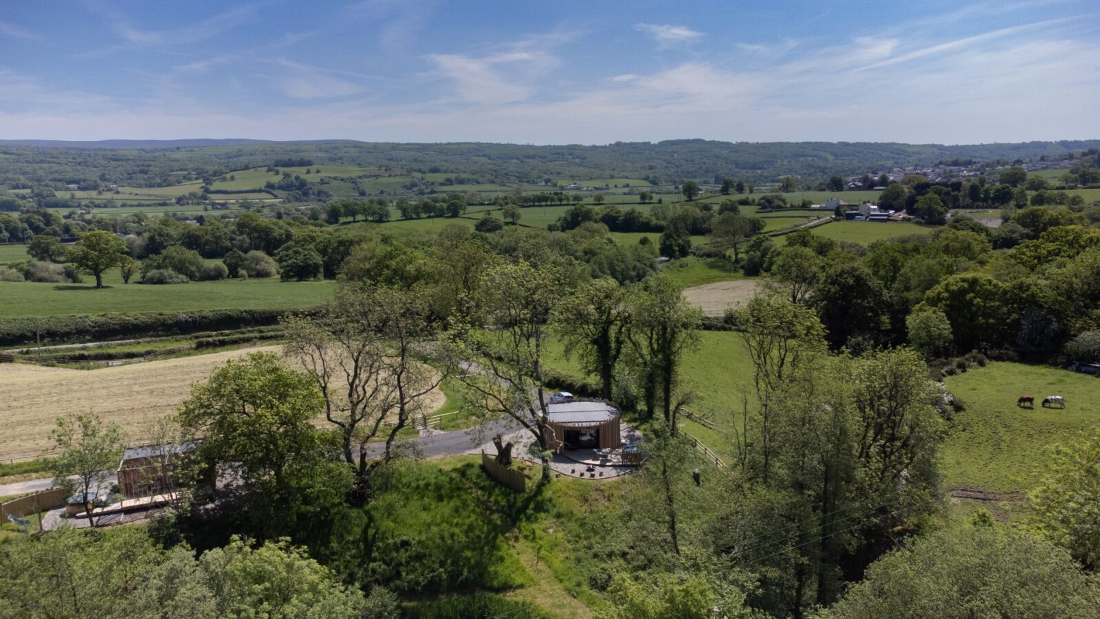 Aerial view of a countryside landscape with green fields, trees, and rolling hills. Pond View Lodges stand in the foreground amid lush greenery. A few scattered houses and grazing animals are visible under a blue sky with wispy clouds, enhancing the serene atmosphere.