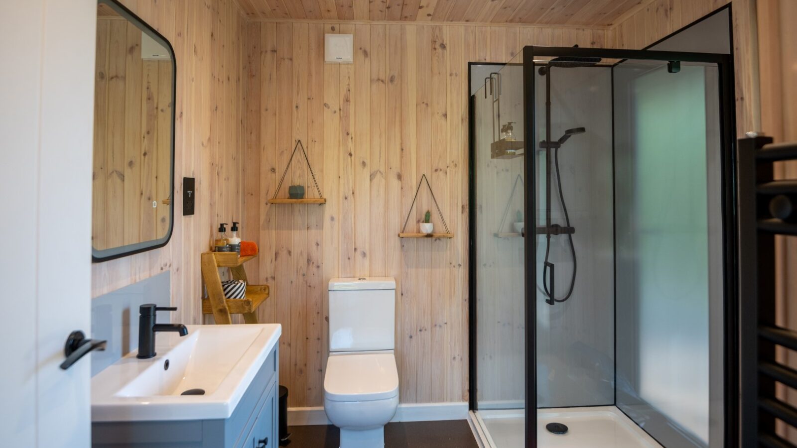 A modern bathroom at Pond View Lodges features wood-paneled walls, a glass-enclosed shower, a white toilet, and a sleek white sink with a black faucet. Shelves display decor items, while natural light brightens the inviting space.