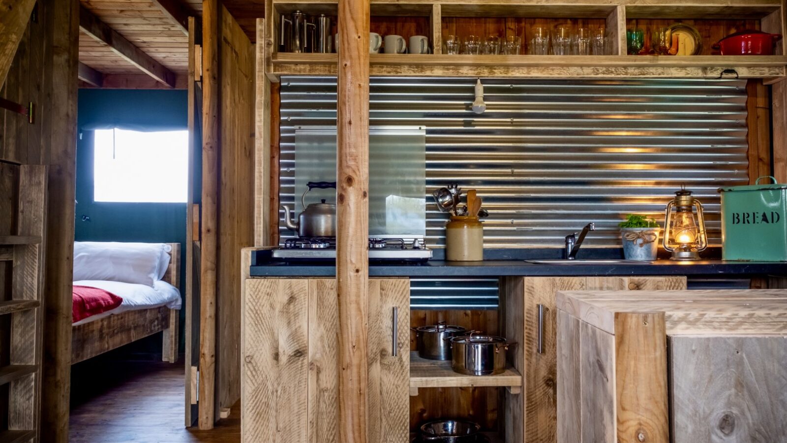 Rustic kitchen at Sloeberry Farm features wooden cabinets, a metal backsplash, and a cozy dining area. A bed with white linens is visible in the adjacent room, evoking the charm of traditional lodges.