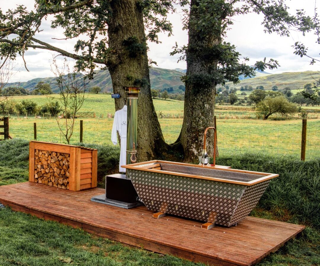 An outdoor bathtub on a wooden deck, flanked by two trees, with a backdrop of grassy fields and hills in The Vale. A wood stack is nearby.