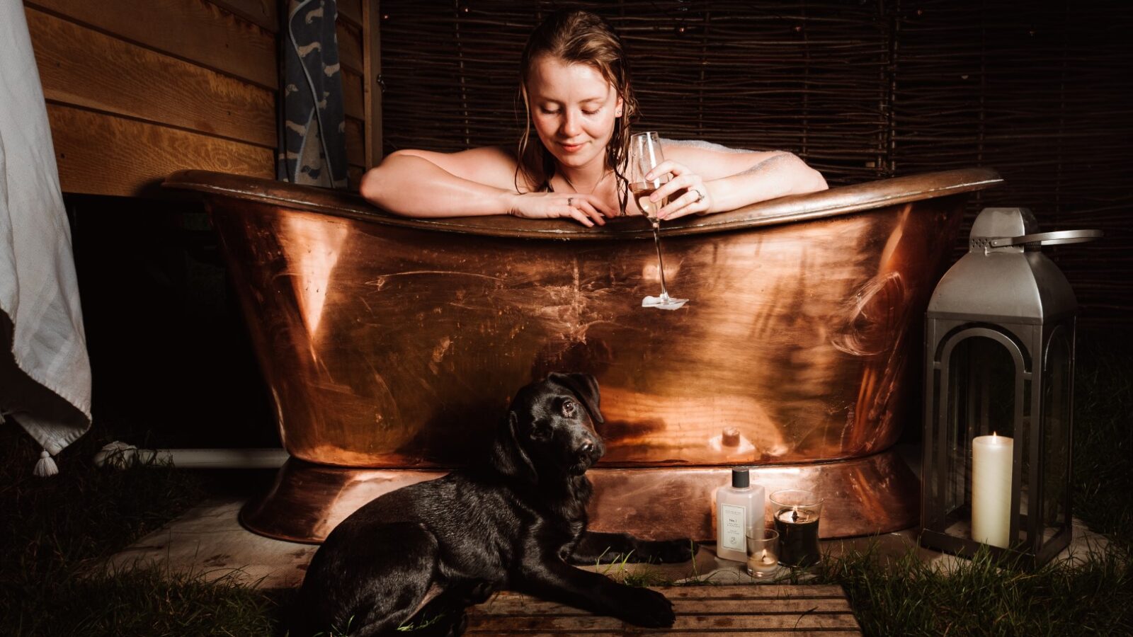 A person relaxes in a copper bathtub outdoors, savoring a glass of wine. Beside the tub, a black dog rests on a wooden mat. Nearby, a lantern with a lit candle and more glasses evoke an aura of serenity—perfect for an Ewe Glamping experience.