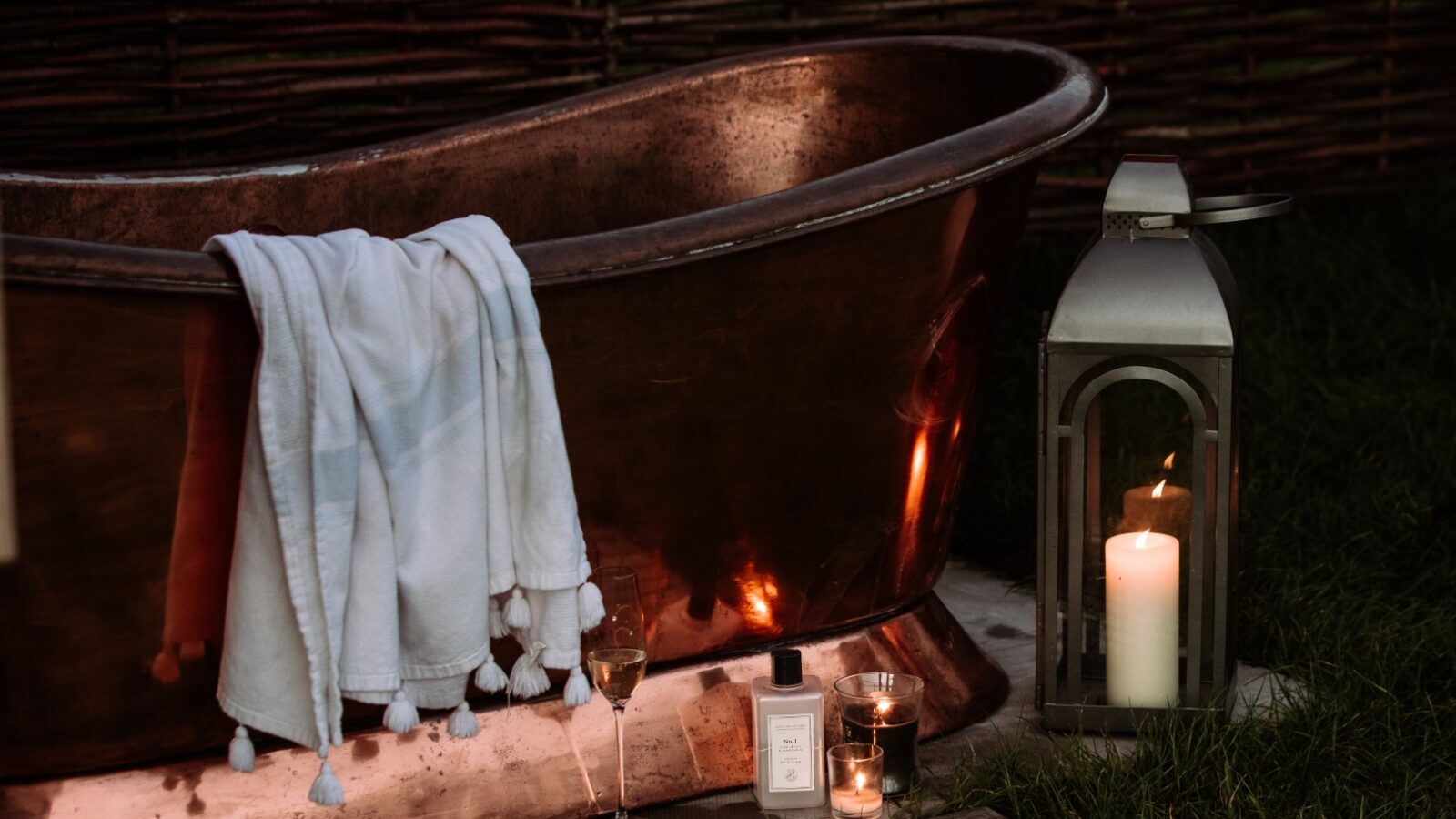 A rustic metal bathtub sits outdoors with a white towel draped over the side, epitomizing Ewe Glamping's charm. A glass of wine, a candle in a lantern, and two small bottles enhance the tranquil vibe. Lush grass and a wooden fence complete this serene Ewe setting.