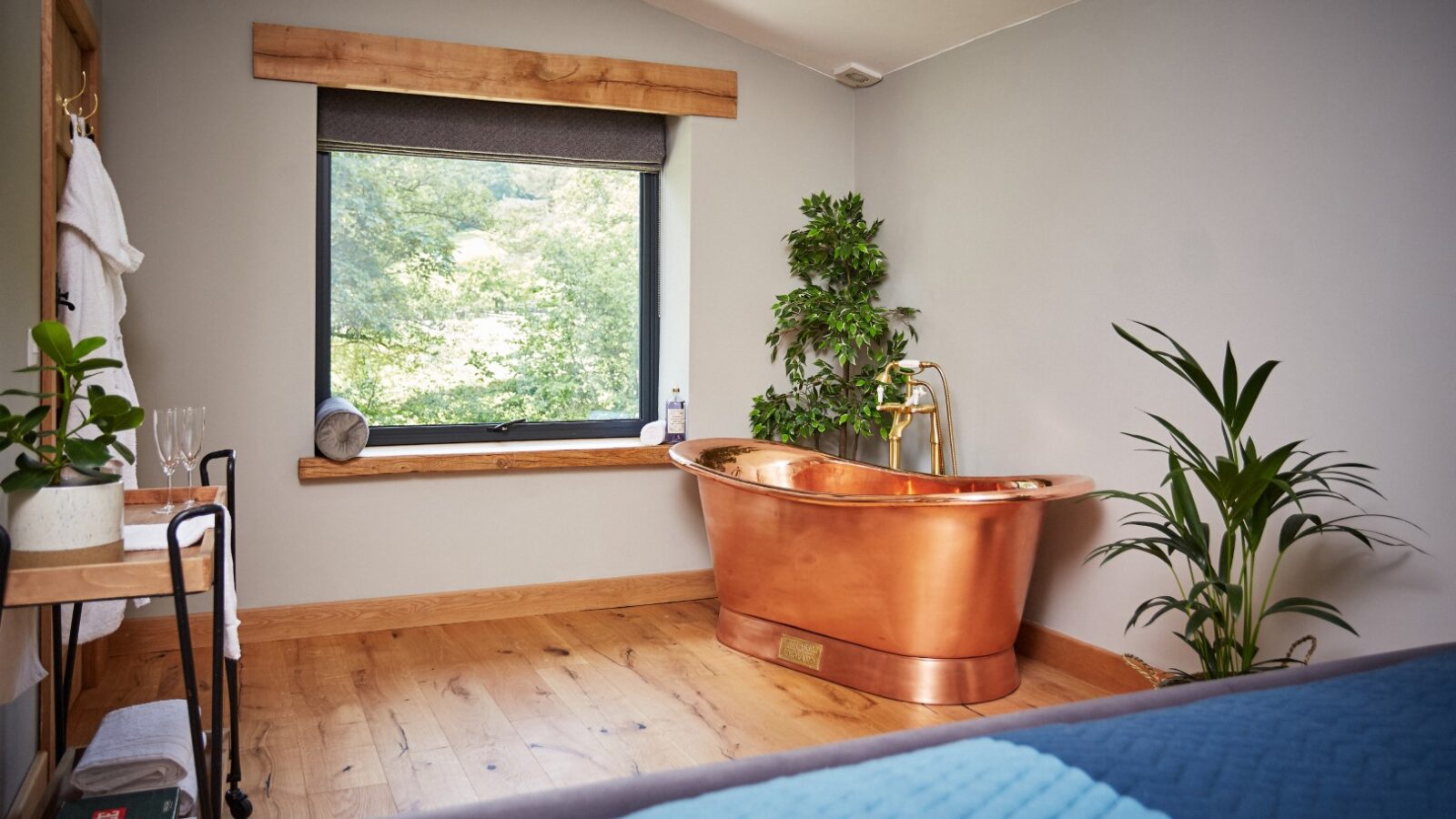 A cozy room by Burrows Dolassey features a copper bathtub beside a large window overlooking lush greenery. The space includes wooden flooring, potted plants, and a towel rack. Neutral walls complement the tranquil atmosphere.