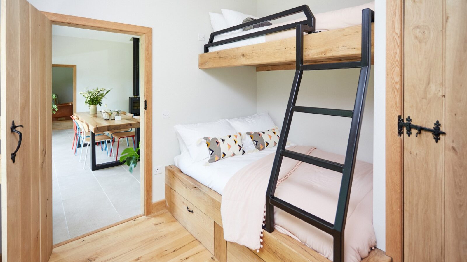 A cozy interior featuring a wooden bunk bed with white and pink bedding exudes a Dolassey charm. The top bunk showcases a sleek black ladder. Wooden floors and walls add warmth, while an open door reveals a dining area with a table and chairs, inviting guests to linger and enjoy the ambiance.