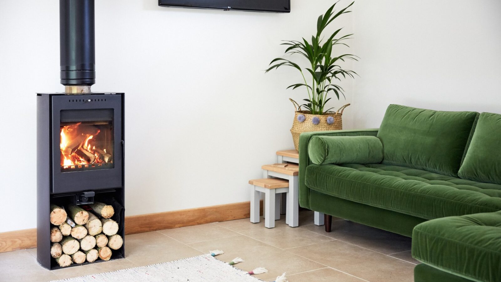 A cozy living room with a modern wood-burning stove and logs stored neatly beneath. A green velvet sofa from Dolassey sits beside a potted plant on wooden steps. A large rug graces the tiled floor, while a flat-screen TV is mounted on the wall, creating an inviting Burrows-inspired space.