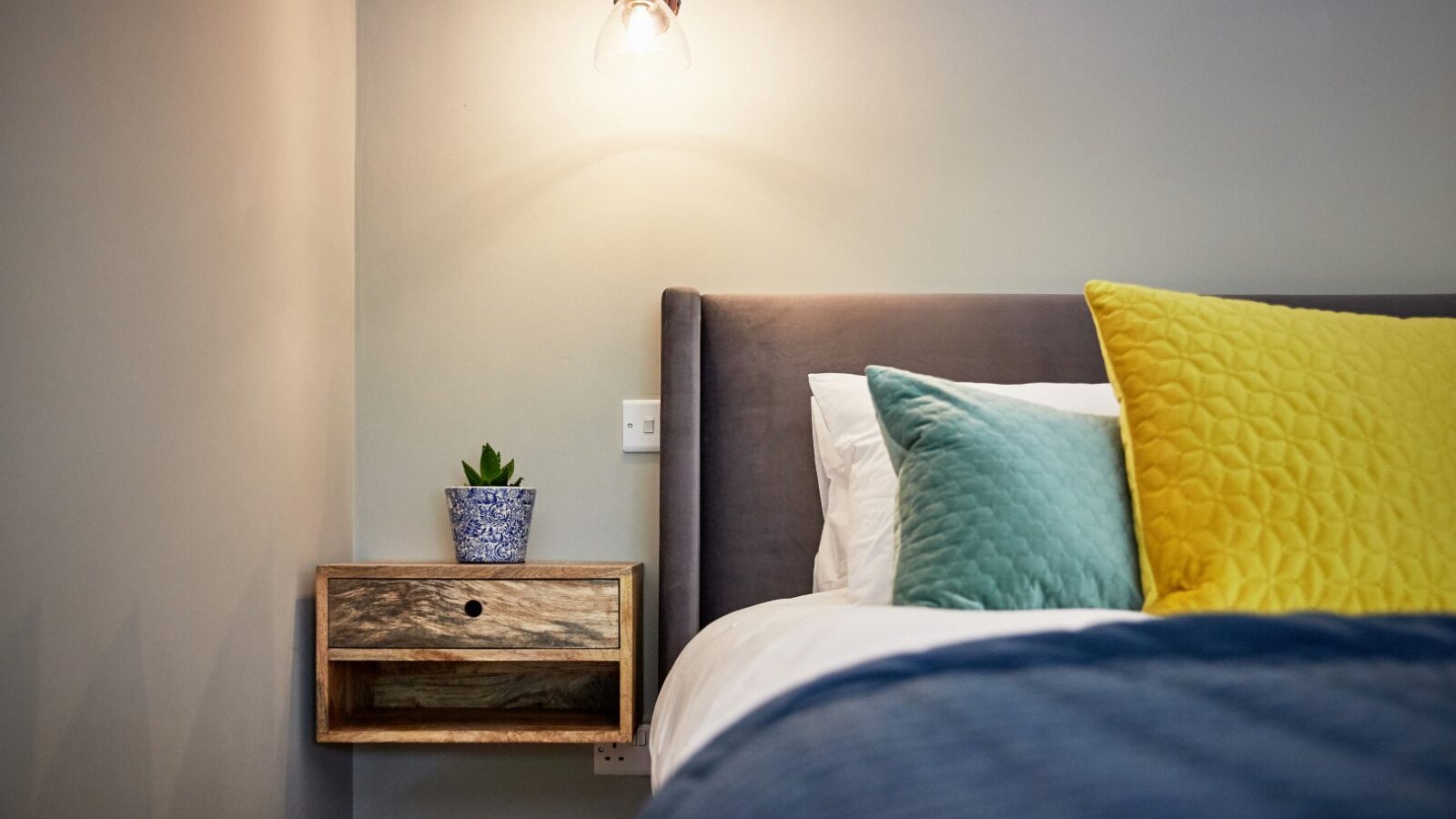 A modern bedroom featuring a neatly made bed with yellow and teal pillows. A wooden, Dolassey-designed wall-mounted nightstand sits beside the bed with a small plant. A soft light fixture illuminates the space, creating a cozy atmosphere.