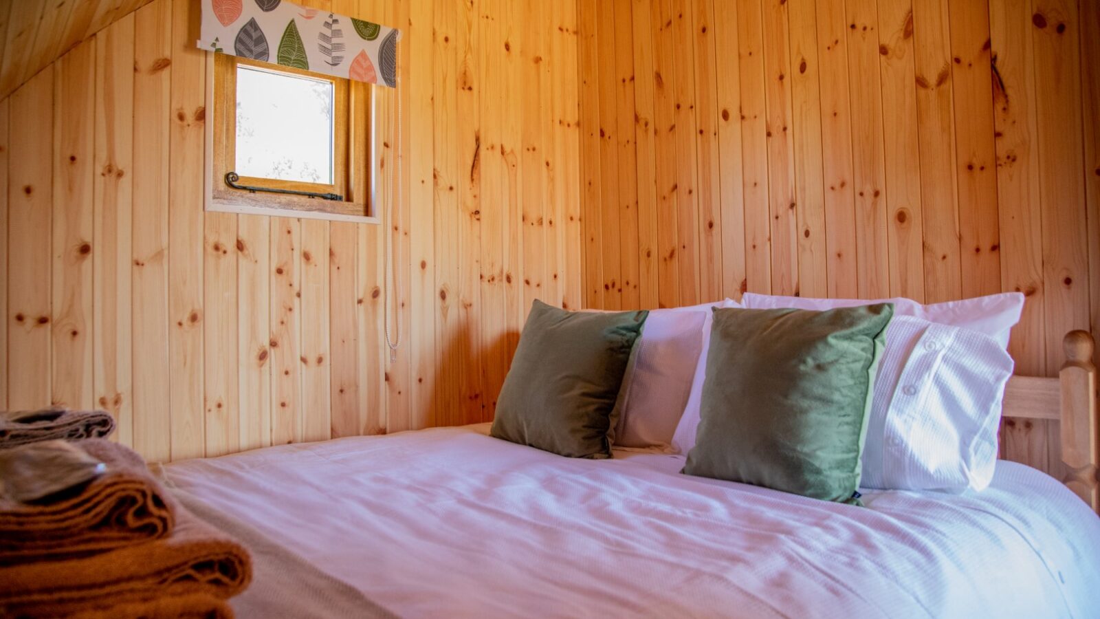 A cozy bedroom with wooden panel walls features a neatly made bed with white linens, green pillows, and a white pillow—perfect for a glamping experience. A small window with a colorful leaf-patterned valance lets in light. Two folded blankets are placed on the bed's corner.