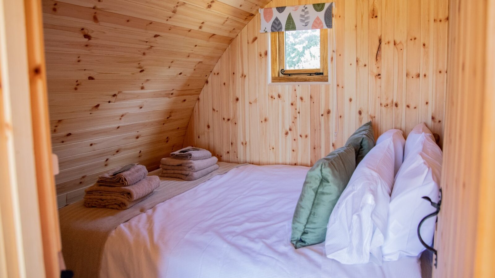 A cozy Barnhorn Glamping bedroom interior with wooden walls and an arched ceiling. There is a neatly made bed with white sheets, green and white pillows, and folded towels placed on the bed. A small window with a colorful valance brings light into the room.