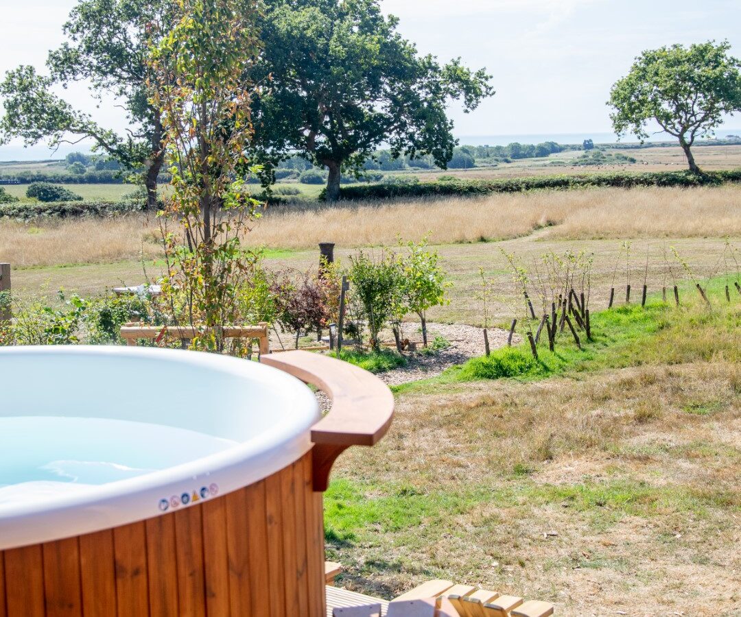 A round wooden hot tub with steps sits on a deck, overlooking a grassy field and a few scattered trees. The sky is clear with a few clouds. This tranquil rural setting, perfect for glamping, suggests relaxation and open space. Experience the charm of Barnhorn in serene comfort.