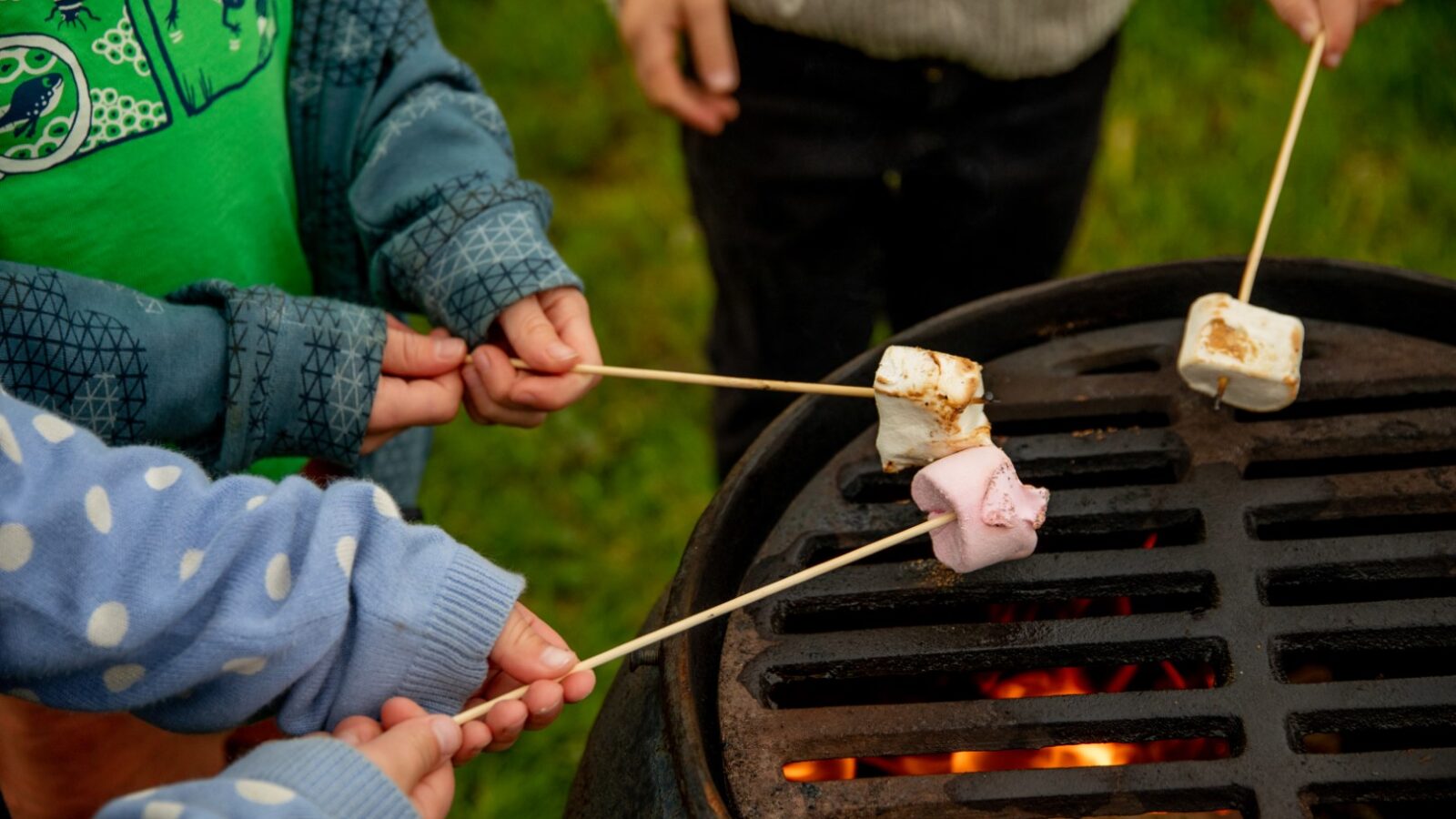 Several children wearing sweaters roast marshmallows over a fire pit at Barnhorn. The marshmallows are on skewers, held close to the flames for toasting. The glamping setting appears to be outdoors, on a grassy area.