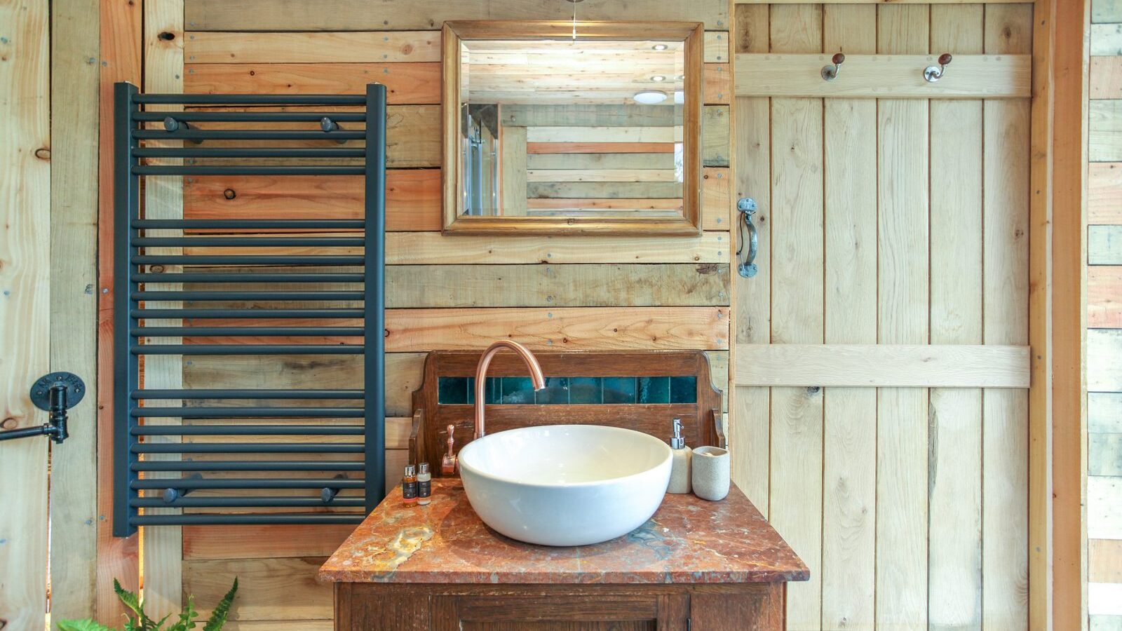 Nestled in a treehouse rental, this rustic bathroom features a wooden interior with a vessel sink on a wooden vanity. A mirror and towel rack complement the space, while a potted plant adds a touch of nature. Experience unique accommodation at Trawscwm Treehouse.