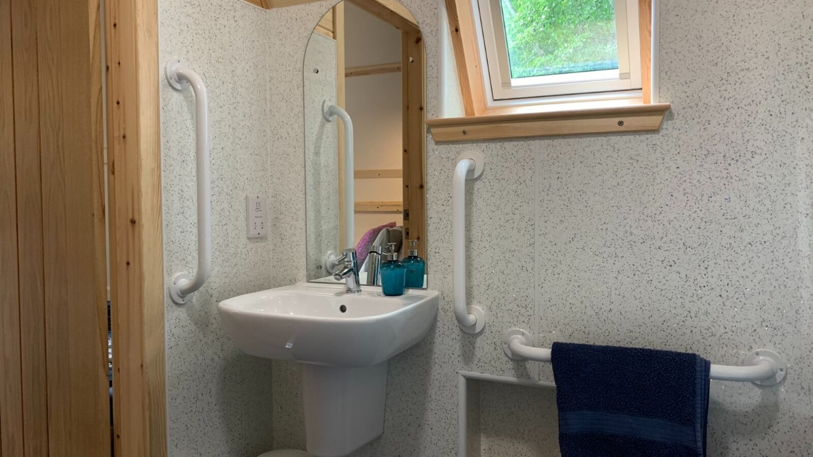 A small bathroom at Forcett Grange features a white sink with two support bars on either side. Above the sink is a mirror, and to its right, a small window. The wall-mounted sink holds two teal cups and soap. A dark towel with wooden accents hangs on a rail beside the sink.