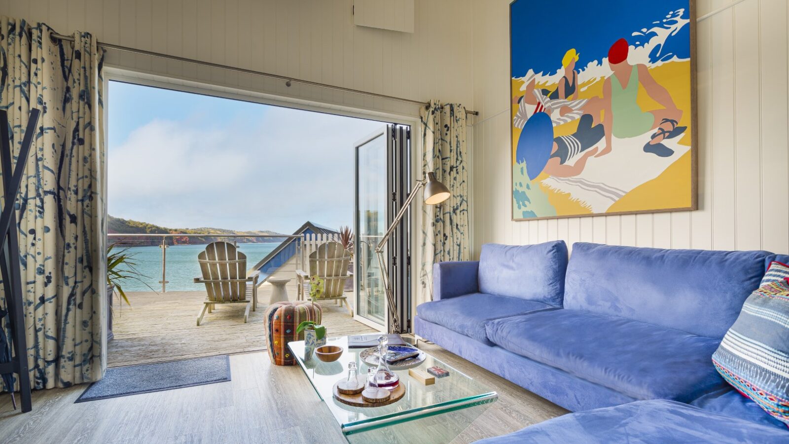 Cozy living room in our beach hut suite featuring a blue sofa, glass coffee table, and vibrant artwork. Open the doors to step onto the deck and soak in the ocean view at Cary Arms.