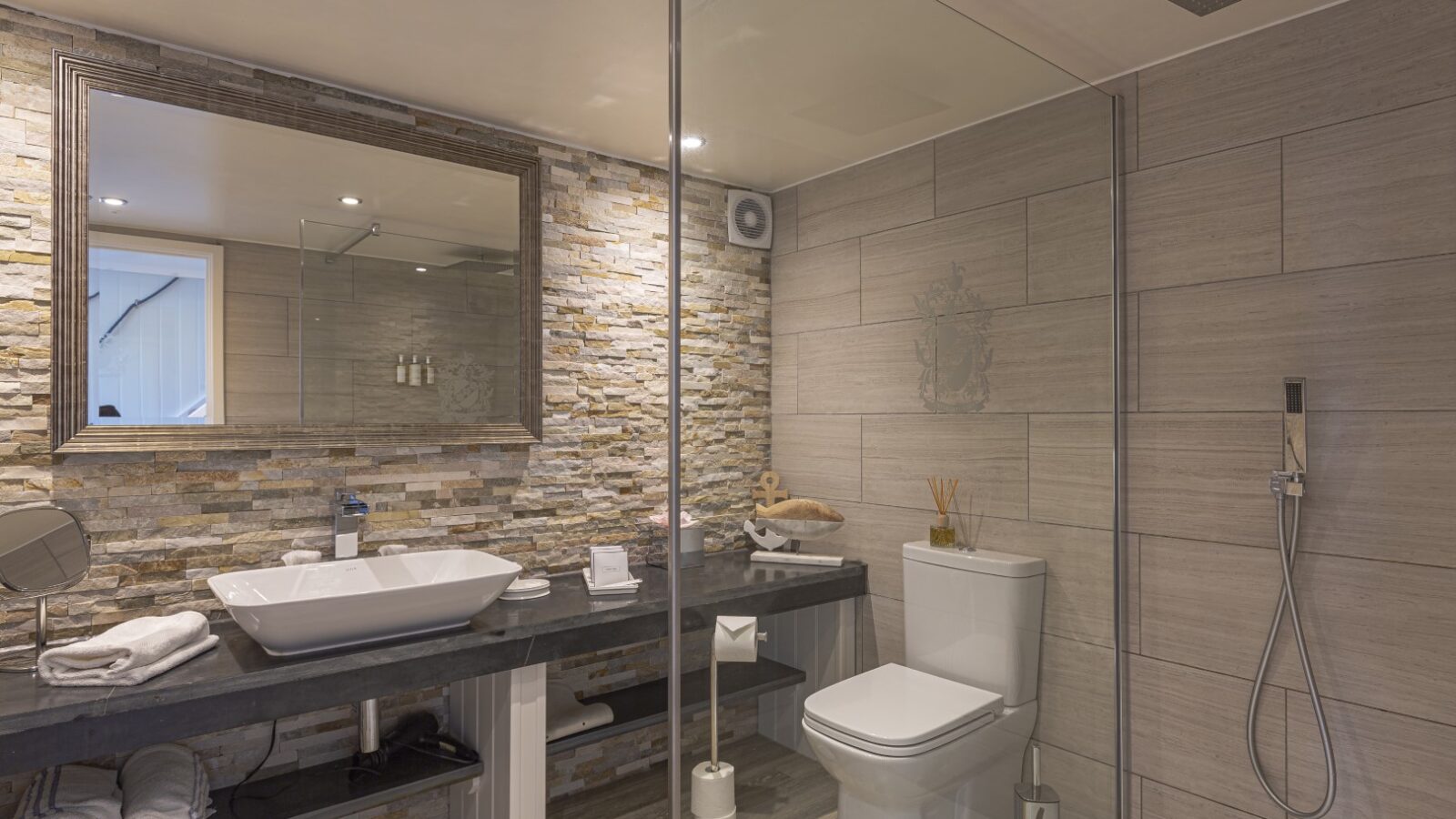 Modern bathroom in our Cary Arms suites, featuring a glass shower enclosure, stone-texture wall, vessel sink, large mirror, and sleek toilet.