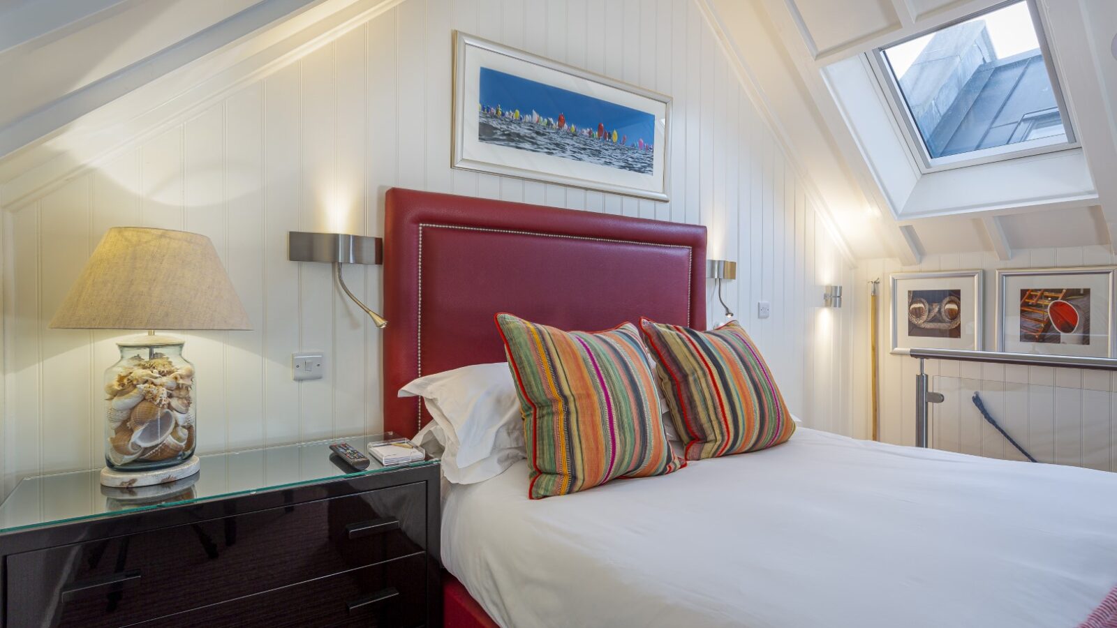 Cozy attic bedroom suite with a red-upholstered bed, striped pillows beside a glass nightstand, and an angled skylight window reminiscent of the charming decor at Cary Arms’ beach huts.