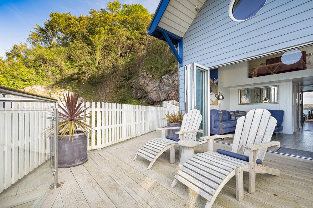 Wooden deck with two Adirondack chairs, a nod to the comfort of Cary Arms suites, alongside a potted plant and a view of a blue seaside house. A white fence and lush greenery provide the backdrop, reminiscent of charming beach huts.