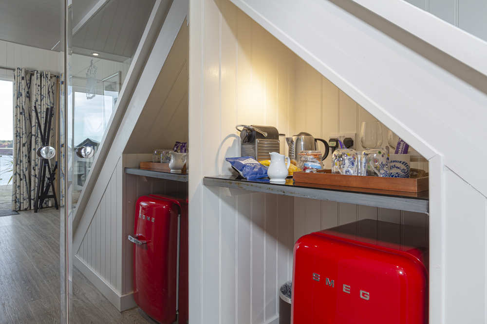 Cozy attic kitchenette akin to charming beach huts, featuring a red fridge, coffee maker, kettle, and tray with cups under a slanted white ceiling. Perfectly complements the style of Cary Arms suites.