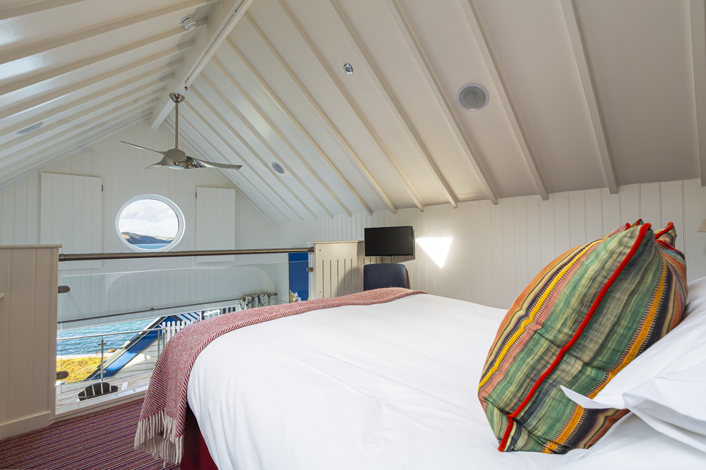 Loft bedroom with a sloped ceiling, round window, and white walls adorned with a bed of colorful pillows. This charming suite overlooks a scenic view reminiscent of idyllic beach huts, offering a tranquil retreat akin to the serene ambiance of Cary Arms.