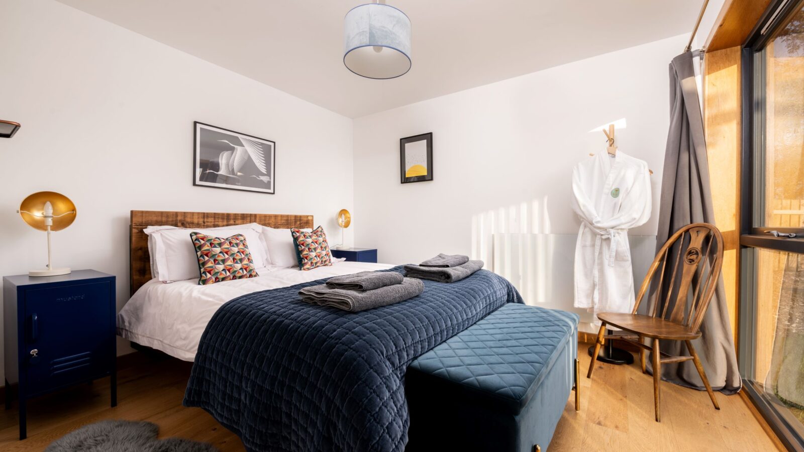 A neatly arranged bedroom evokes a cozy cabin vibe, featuring a double bed with blue bedding, framed artwork, and a robe on a coat rack by the window.