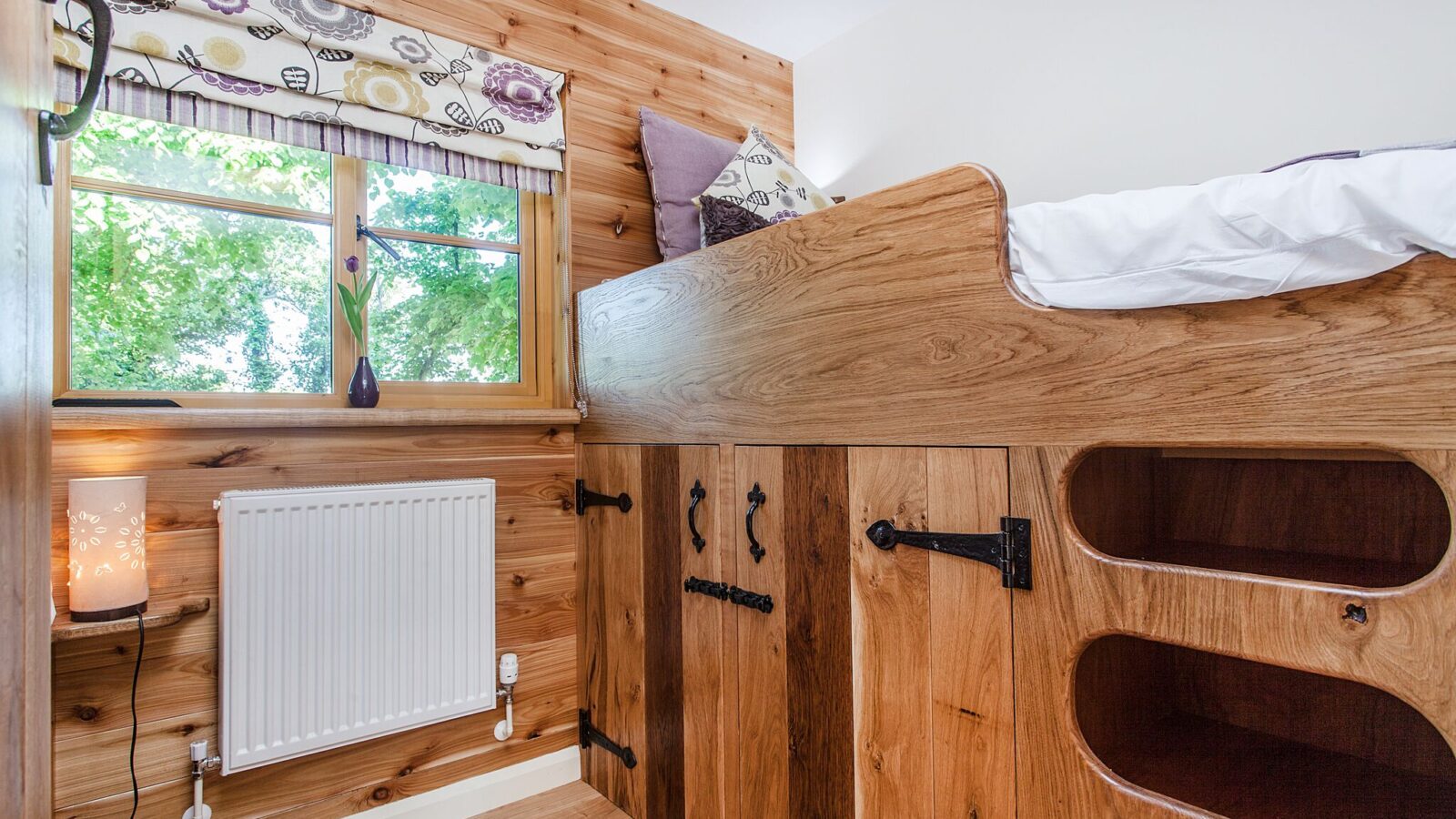 Wood-paneled room with a raised bed, storage cabinets, a radiator, and a window with floral curtains overlooking trees, reminiscent of the charming retreats at Lavender Hill Treehouses.