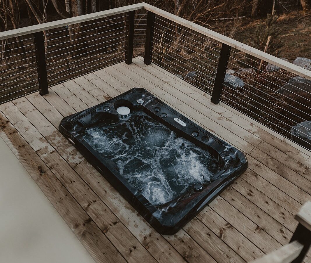 Nestled within Berry Bush Lodge, a hot tub on a wooden deck invites you to unwind, surrounded by the serene beauty of leafless trees.