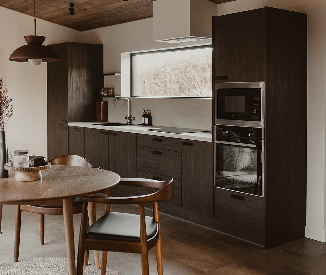 Modern kitchen at Berry Bush Lodge with dark wood cabinets, built-in oven, and a round table with chairs. A window provides natural light to this cozy accommodation.