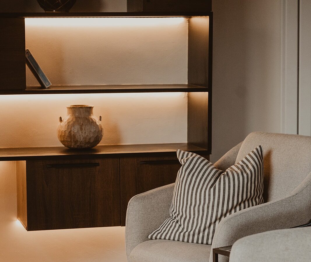 The Berry Bush Lodge-style living room features a beige armchair, a striped pillow, wall-mounted shelves, a decorative vase, and warm lighting that creates a cozy yet modern ambiance.