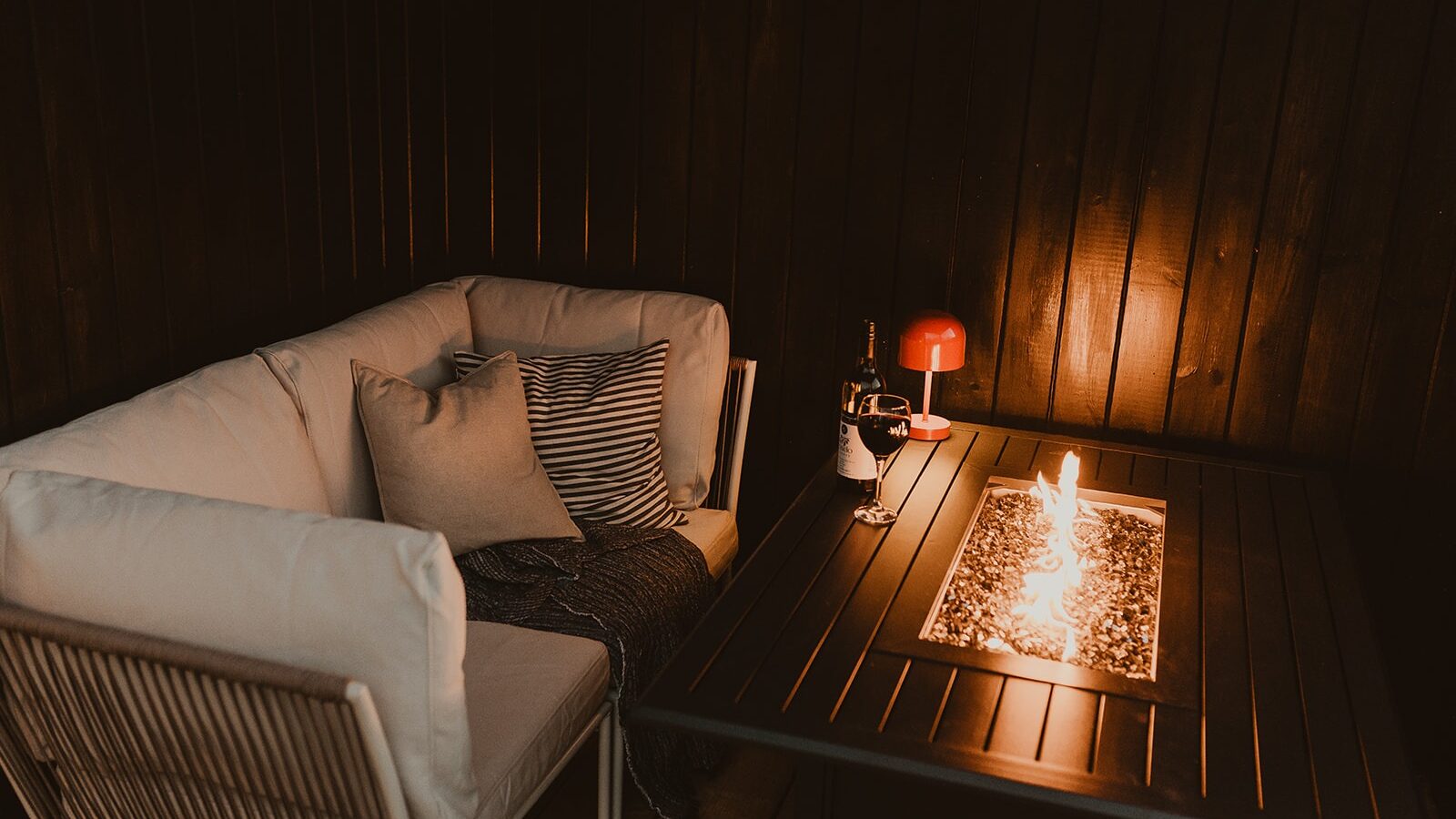 Cozy corner at Berry Bush Lodge with a couch, pillows, and a lit fireplace table. A red lamp and wine glasses complete the warm atmosphere.