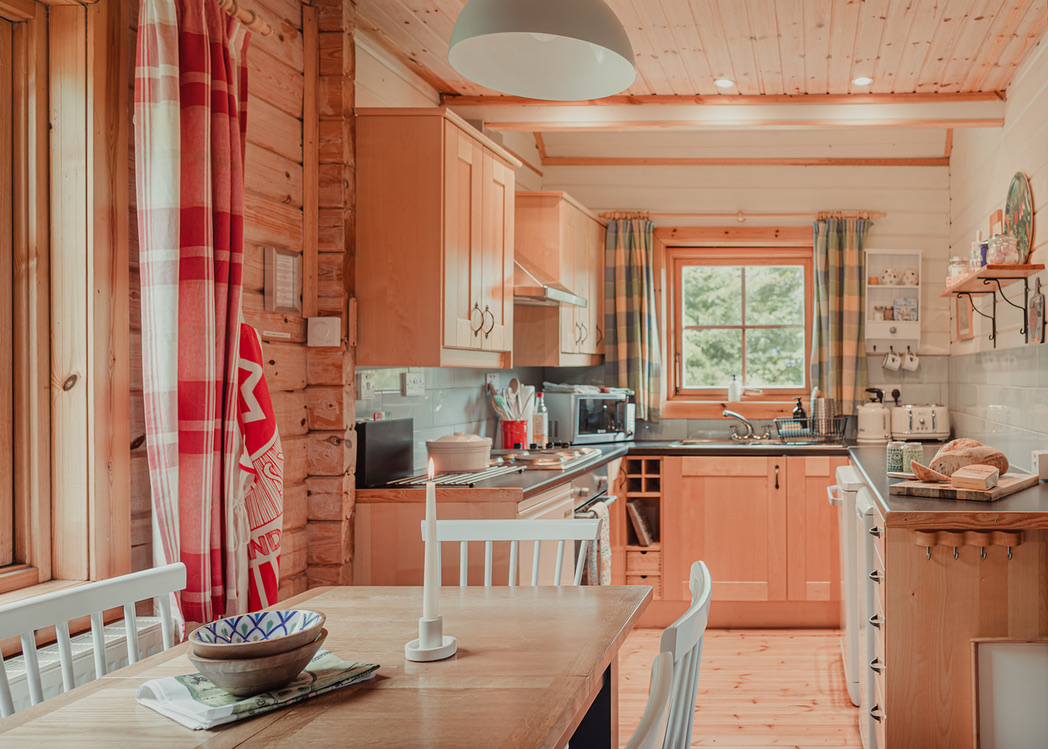 A cozy cabin kitchen at Big Sky Lodges with wooden walls and ceiling features red and blue-checkered curtains, light wooden cabinets, and white countertops. A wooden dining table with white chairs sits in the foreground, and a window above the sink offers a view of green trees.