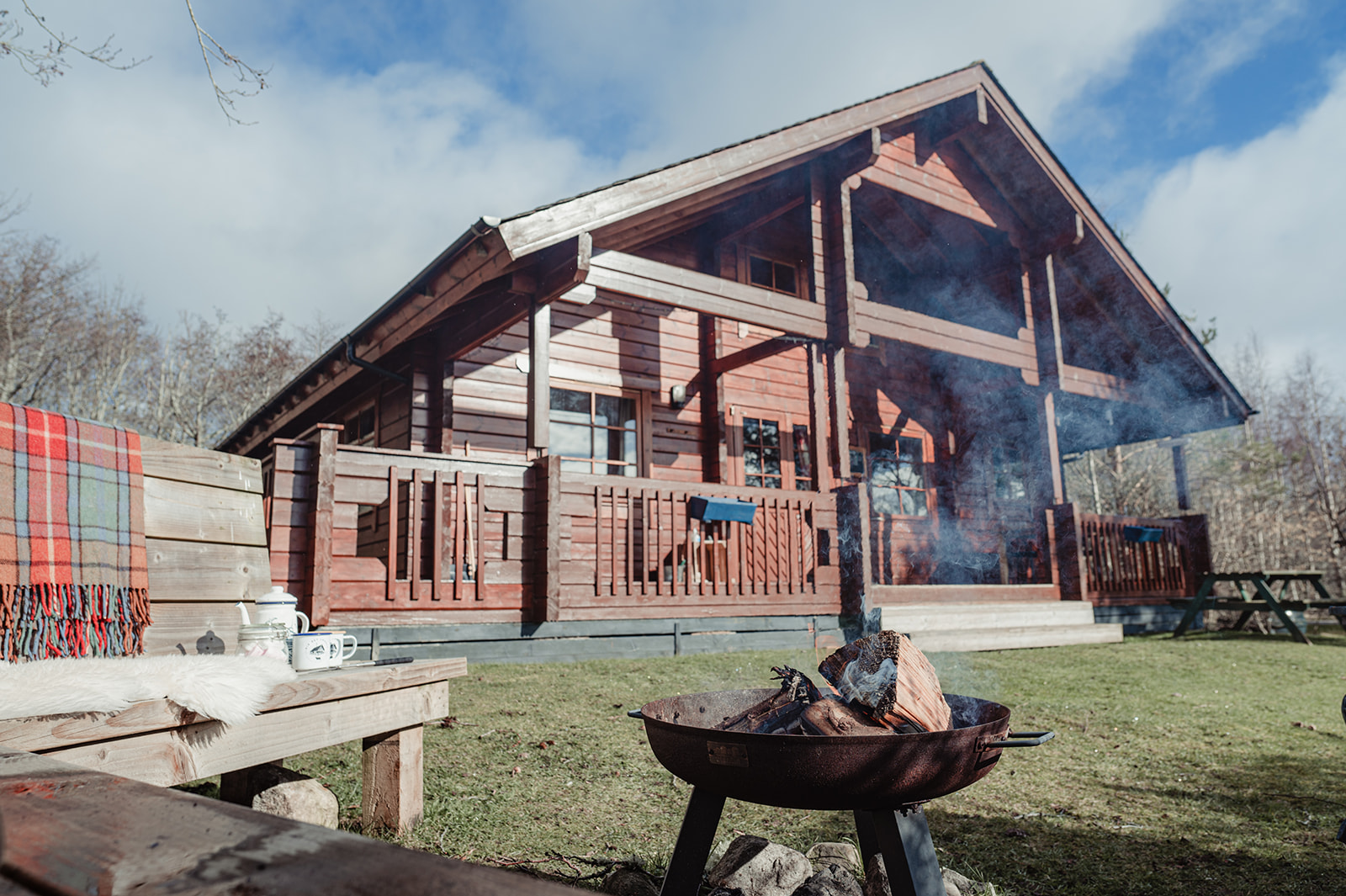 A wooden cabin with a large porch sits in a scenic outdoor setting at Big Sky Lodges. In the foreground, there's a fire pit with burning logs, a wooden bench adorned with a red plaid blanket, and a small white coffee maker. The sky is partly cloudy.