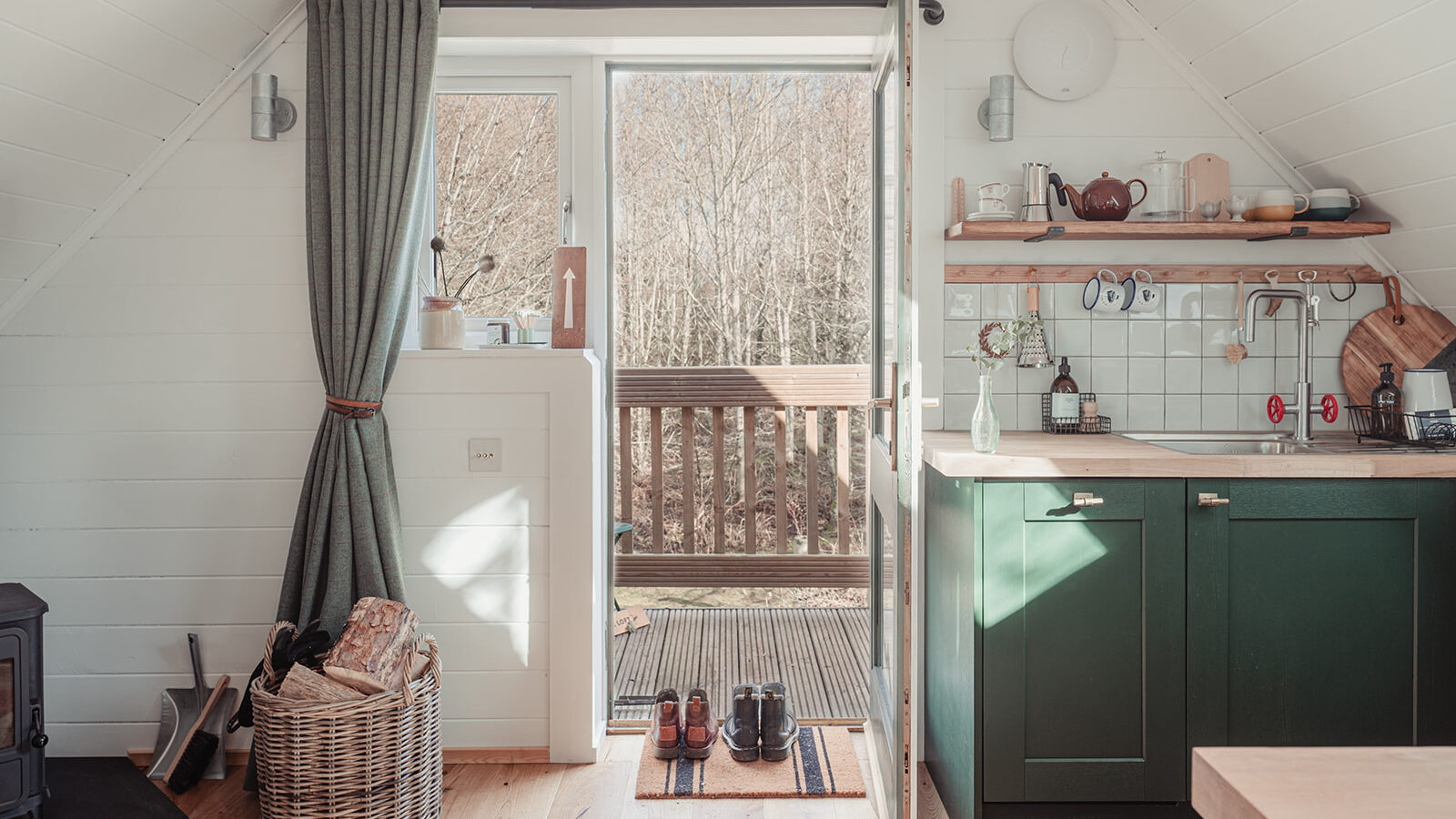 A cozy, light-filled cabin at Big Sky Lodges features a green kitchen counter, utensils, and a stove in the corner. An open door reveals a deck with a forest view. A curtain is tied back, a basket near the stove holds firewood, and two pairs of shoes are placed near a small mat.