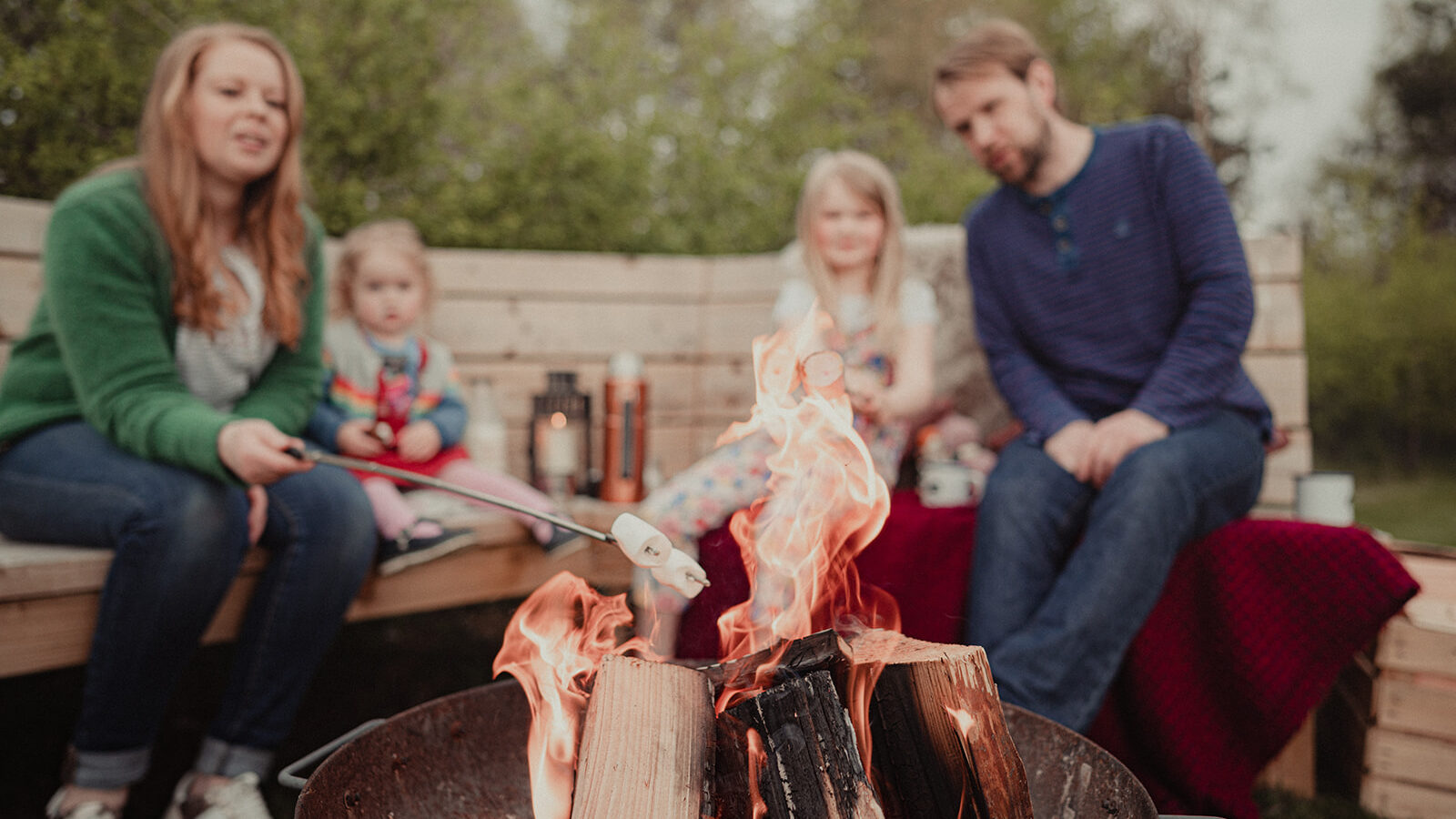 A family of four sits around a fire pit at Big Sky Lodges, roasting marshmallows. The woman on the left, wearing a green jacket, holds a marshmallow over the fire. Two children, one in colorful clothes and another in a gray outfit, and a man in a blue shirt are also seated. Trees are in the background.