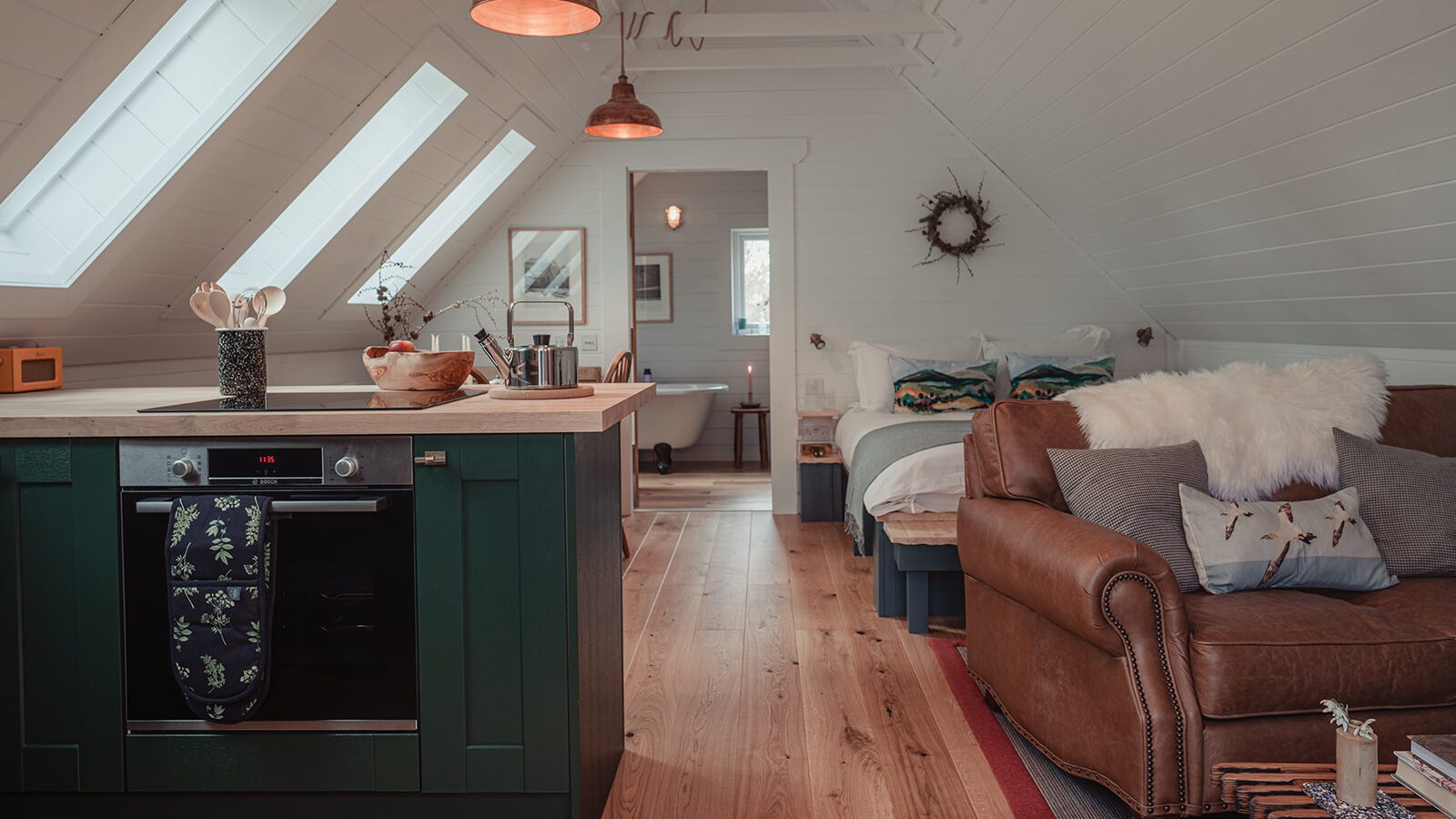 A cozy attic studio by Big Sky Lodges with wooden floors, featuring a kitchen area with green cabinets, a small island, and modern appliances. To the right, there's a brown leather sofa, a bed with white linens, and rustic decorations. Skylights and pendant lights brighten the space.