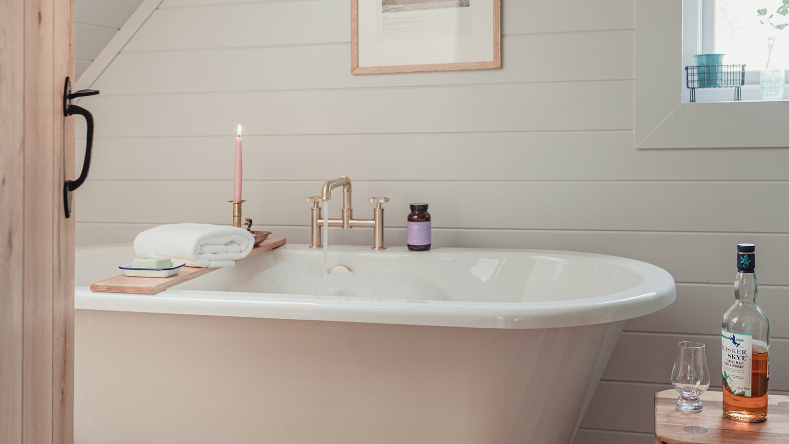 A cozy bathroom with a white clawfoot bathtub against a paneled wall, reminiscent of rustic lodges. The tub is adorned with a wooden bath tray holding a candle, towel, and toiletries. A framed picture of the Big Sky hangs above, and a small windowsill features a plant. A stool with a bottle and glass sits nearby.