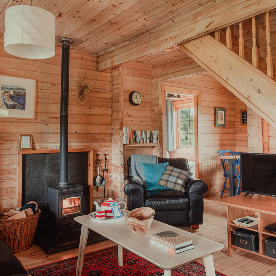 A cozy wooden cabin living room at Big Sky Lodges features a lit wood stove, a black armchair with colorful cushions, and a wooden staircase. A coffee table with books and tea set rests on a red patterned rug. Shelves with books, a framed picture, and a TV adorn the charming space.