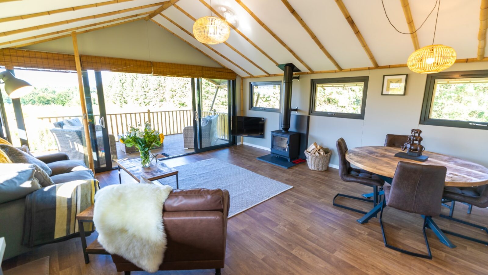 A cozy Birdholme living room with wooden flooring, large windows, a wood stove, a brown leather chair, and a round dining table offers a perfect glamping vibe.