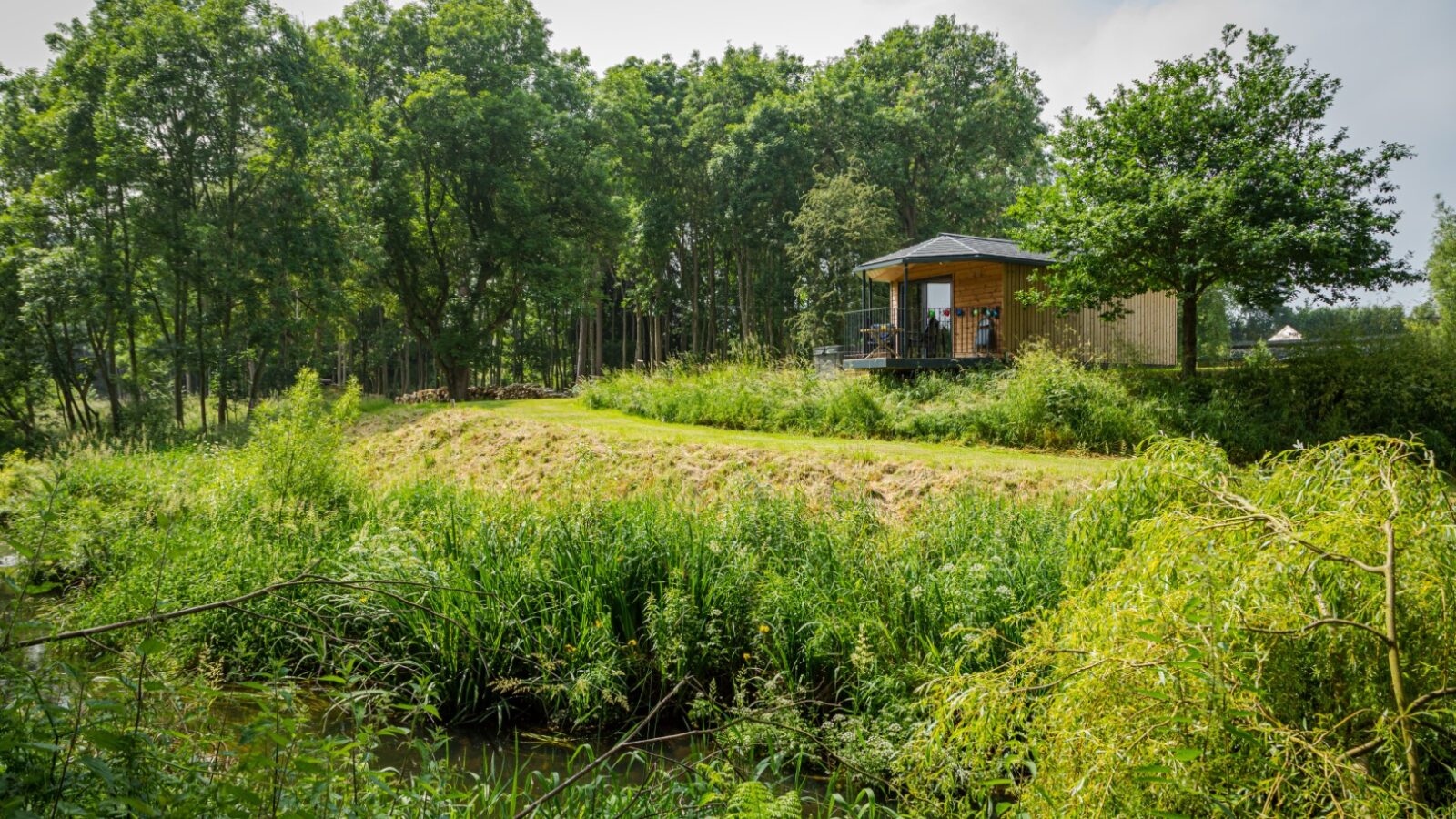 A small wooden cabin sits among trees beside a grassy area and a stream, reflecting its riverside charm under a cloudy sky.