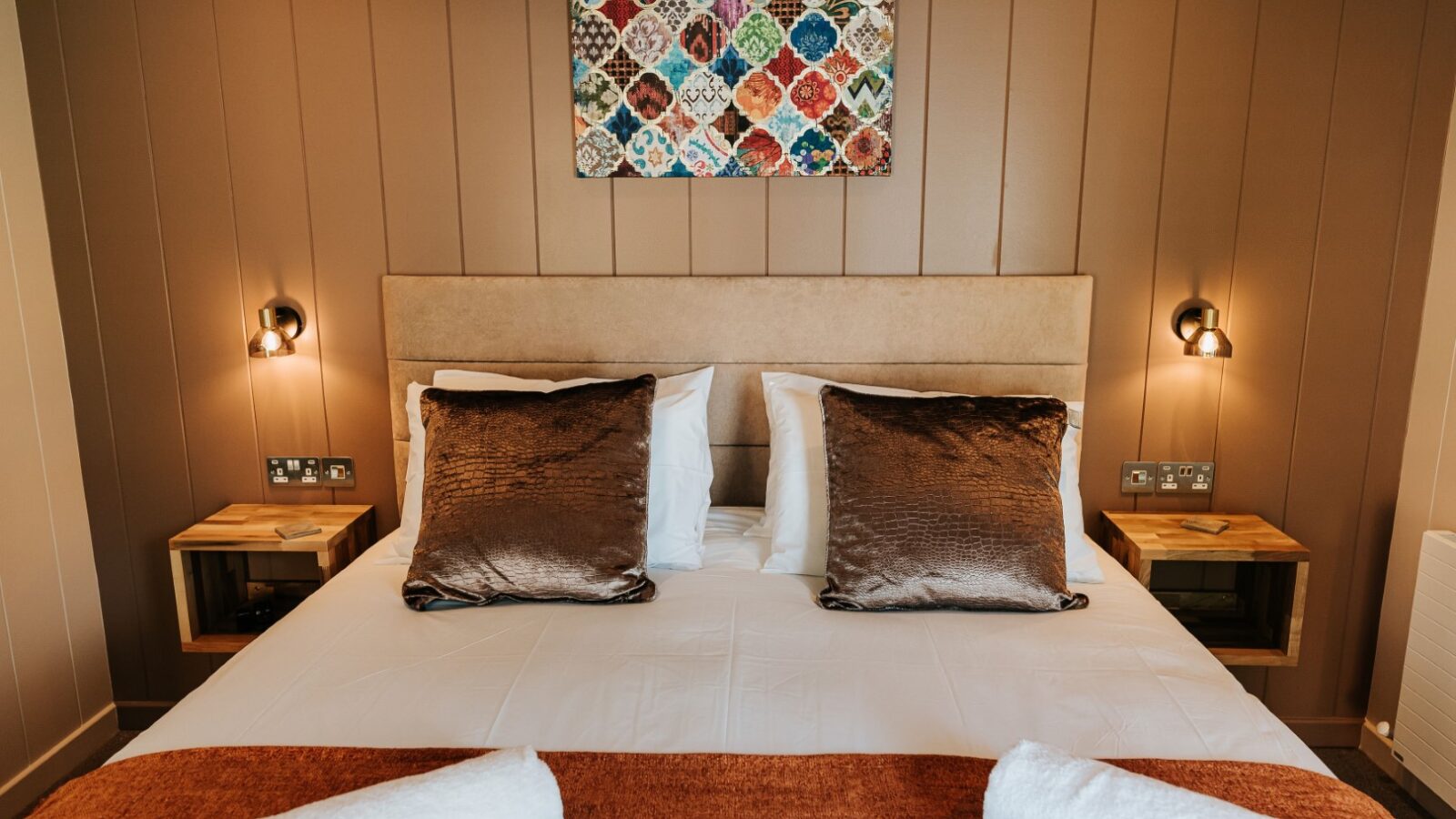 A neatly made bed with brown pillows, white sheets, and towels in a cozy room at Bowland Retreat Lodges features wall lamps and a colorful quilted artwork.