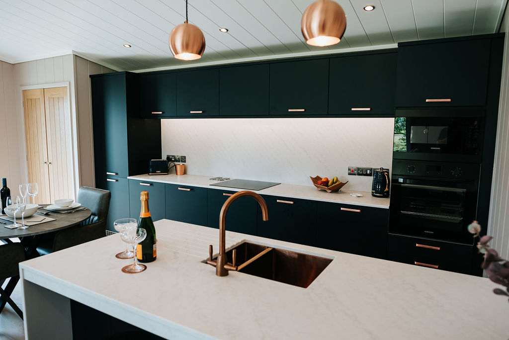 Discover a modern kitchen at Bowland Retreat, featuring black cabinets, white countertops, copper fixtures, and elegant hanging lights. This stylish space is perfect for dining at a set table after exploring nearby lodges.