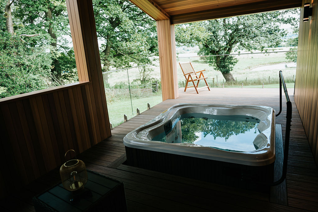 Experience tranquility at Bowland Retreat, where the covered deck features a hot tub and a wooden chair overlooking a grassy field through glass barriers. This idyllic setting, nestled among the trees, makes for the perfect escape at one of our charming lodges.