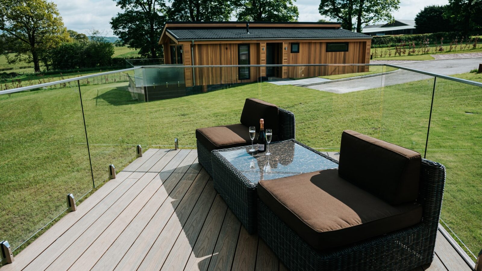 The wooden deck at Bowland Retreat features a glass railing, two brown wicker chairs, and a glass table with wine glasses, all overlooking the serene garden and charming wooden lodges.