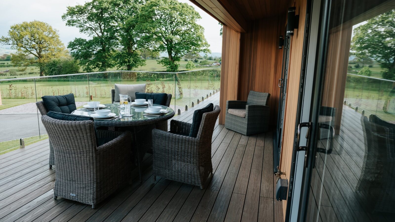 Nestled within Bowland Retreat, a deck with a round table set for six is surrounded by wicker chairs, overlooking a lush landscape with trees and glass railing.