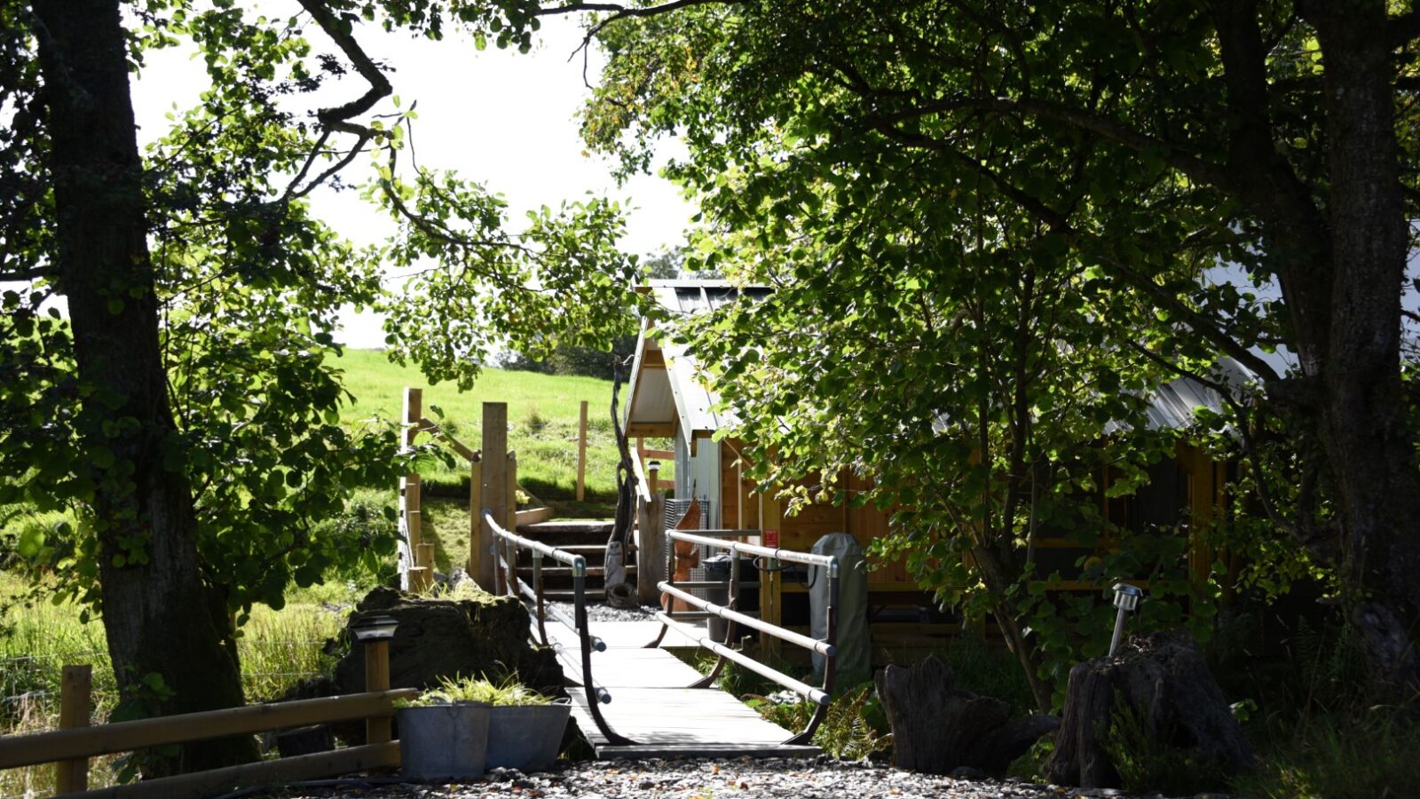 A small wooden bridge leads to the rustic Stellar Lodge, surrounded by trees and greenery on a sunny day.