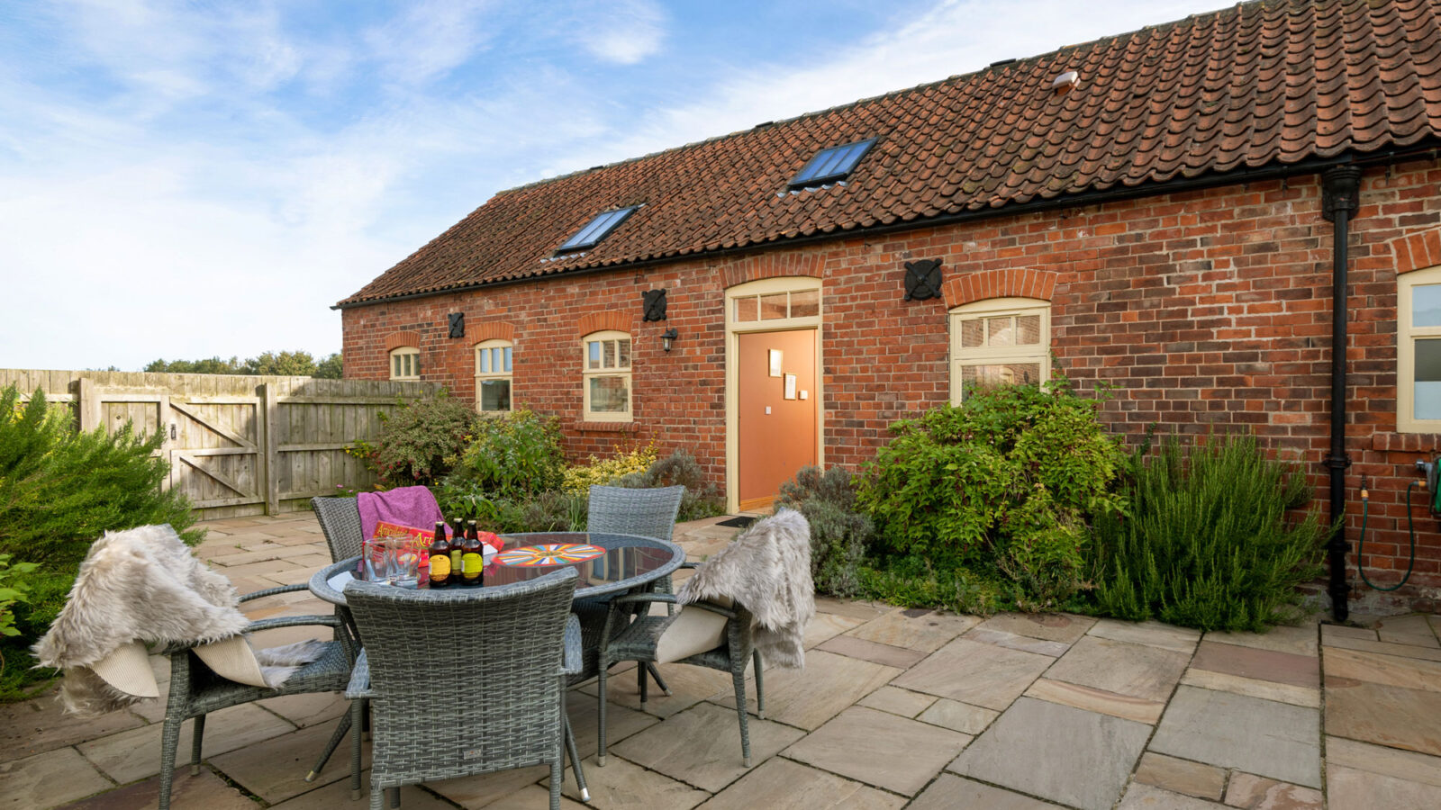 A cozy outdoor patio with a round table and four wicker chairs, each with a sheepskin throw. The table is set with drinks and snacks. The patio, part of the charming Broadgate Farm Cottages, belongs to a brick house with a red-tiled roof and several windows, framed by shrubs and greenery.