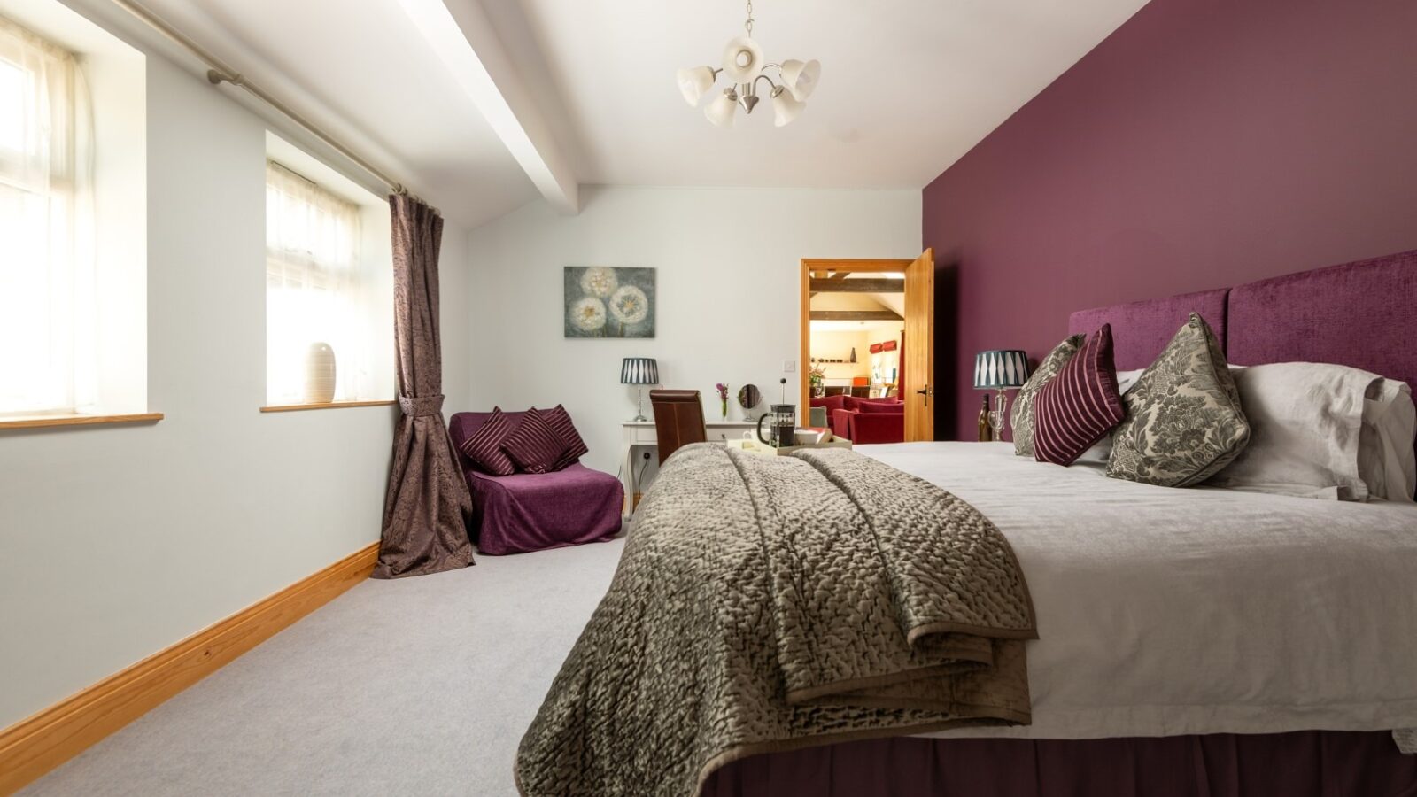 A cozy bedroom at Broadgate Farm with a large bed covered in gray and purple bedding, pillows, and a throw blanket. The walls are a mix of white and purple. There are two windows with light curtains, a small seating area with chairs, and a chandelier hanging from the ceiling.