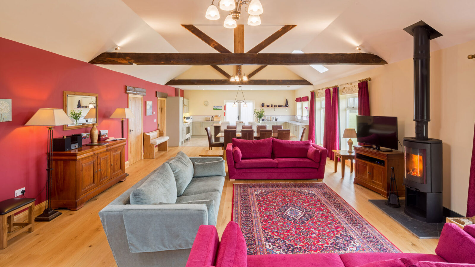 A spacious living room with a vaulted ceiling and exposed wooden beams, reminiscent of the charming style of farm cottages. The room features red and blue sofas, a wood-burning stove, a large patterned rug, wooden furniture, and natural light streaming through windows with red curtains. This inviting area at Broadgate Farm seamlessly opens to the kitchen.