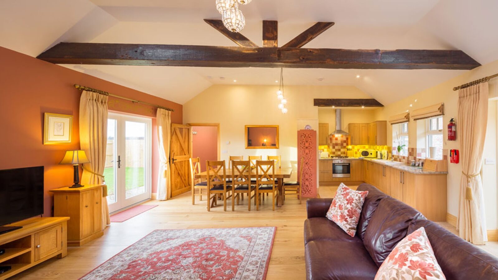 A spacious, well-lit open-concept living room at Broadgate Farm Cottages features a leather sofa with patterned cushions, a large rug, and a mounted TV. The adjoining dining area includes a wooden table and chairs. Modern kitchen appliances and wooden cabinets stand in the background. Exposed wooden beams accentuate the ceiling, and warm tones abound with sunlight streaming in from double glass doors that lead to an