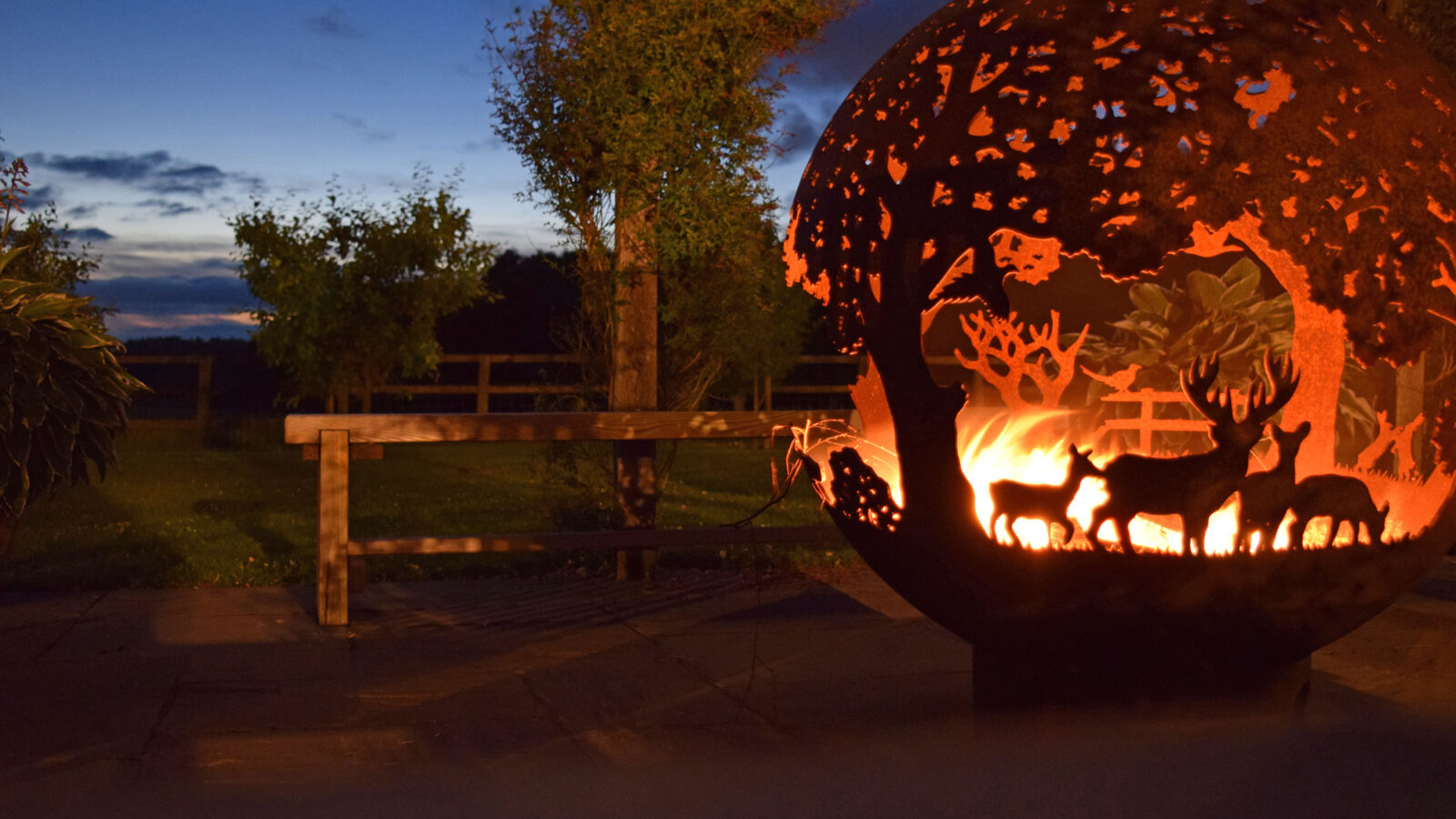 A spherical fire pit with intricate cutout designs of deer and trees glows warmly against the backdrop of a serene outdoor landscape at Broadgate Farm Cottages at dusk. The sky is a deepening blue, and trees and a wooden fence are visible in the dim light.