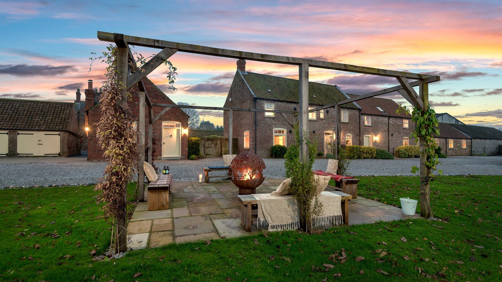 A picturesque outdoor seating area under a wooden pergola with plants, featuring cushioned benches around a modern spherical fire pit. The scene is set on a stone patio at Broadgate Farm Cottages, in front of a rustic brick house during a vibrant sunset. Fallen leaves dot the grass.